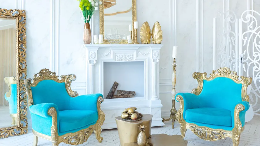 A luxurious white and gold living room featuring two ornate turquoise armchairs with gilded frames flanking a white fireplace mantel adorned with gold leaf decor, a gold mirror, and a vase of green flowers.