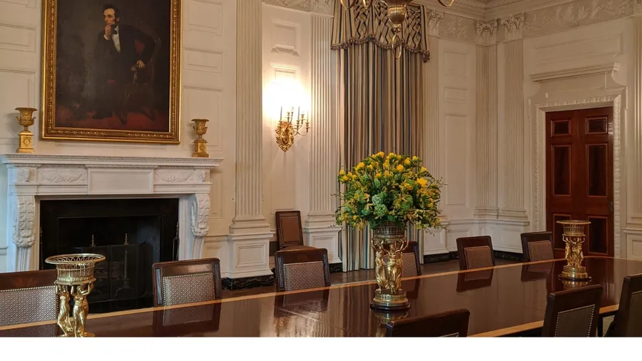 A view of the White House State Dining Room featuring a large wooden table, chairs, a fireplace, and a portrait of Abraham Lincoln above the mantle, with a floral arrangement on the table.