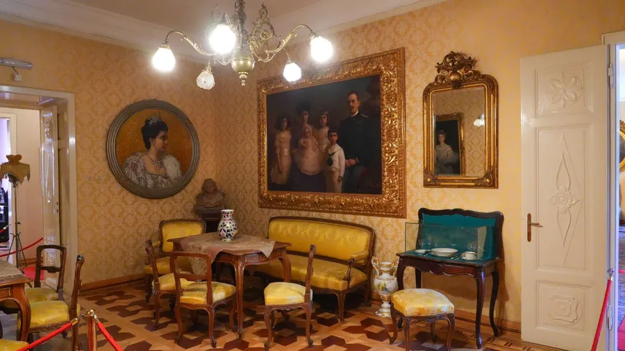 An ornate room, featuring period furniture, framed paintings, and a chandelier.