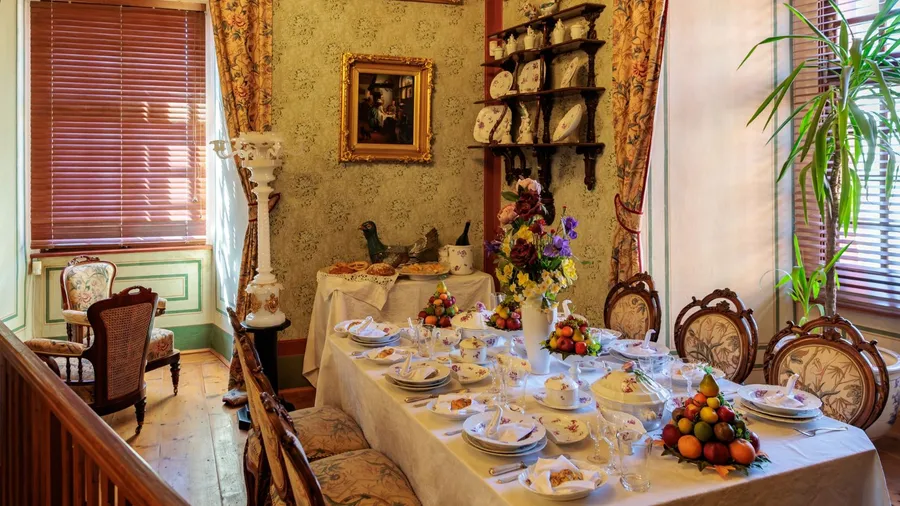 A formal dining room set with a long table laden with dishes and fruit arrangements, featuring ornate furniture, patterned wallpaper, and a large window with wooden blinds.