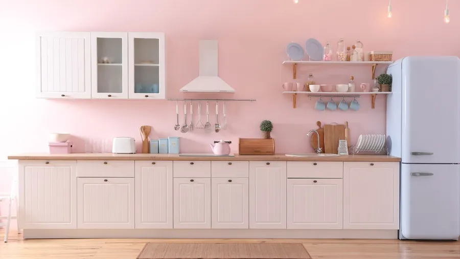 A kitchen interior with warm white cabinets, wooden countertop, blue fridge, floating shelves with cups and jars, and a range hood on the pastel pink wall.