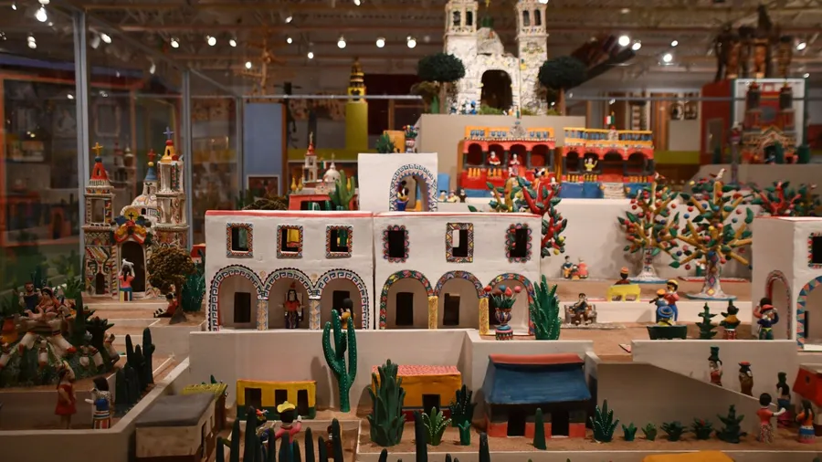 A detailed miniature village scene display, featuring numerous small buildings, figures, and elements like cacti and trees, under bright lighting in what appears to be a museum exhibit.