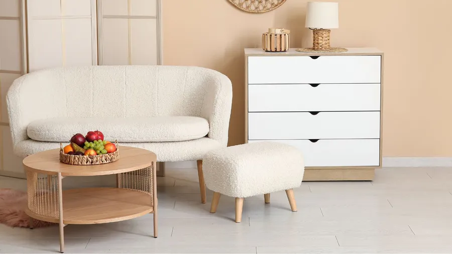 A living room interior with a cozy white sofa, ottoman, console table with lamp, and a coffee table with a fruits basket.