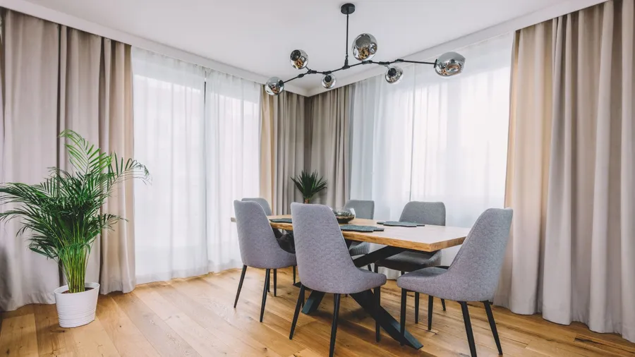 A stylish dining room interior with a wooden table, chairs, plant, curtains, and chandelier hanging from the ceiling.