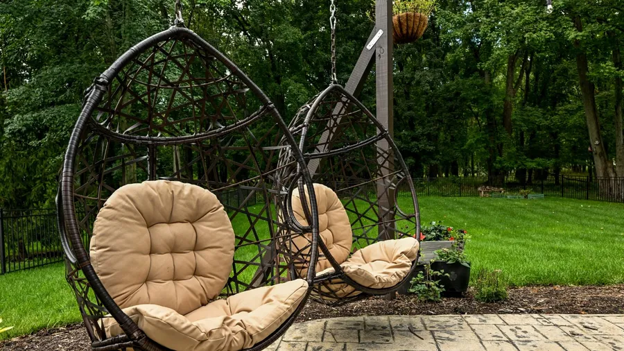 Two hanging wicker egg chairs with beige tufted cushions in a garden.