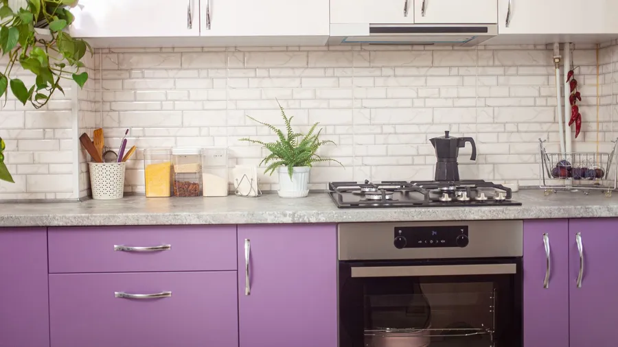 A kitchen interior with white and lavender cabinets, granite countertop with stove, oven, and plants.
