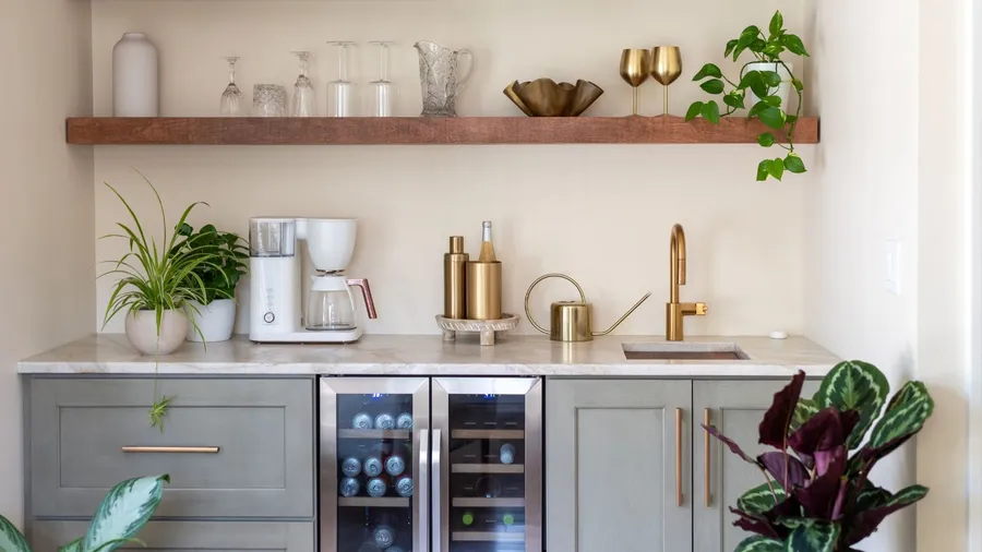 A kitchen interior with marble countertop, decorative metallic accents, and wooden open shelf with glassware, and potted plants.