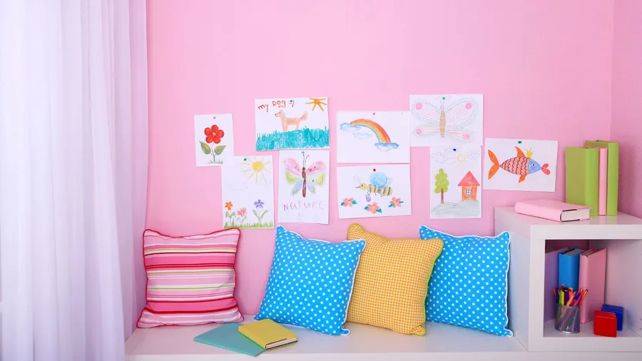 A children's room interior with a white bookshelf, colorful pillows, and kid's artwork on a pink wall.