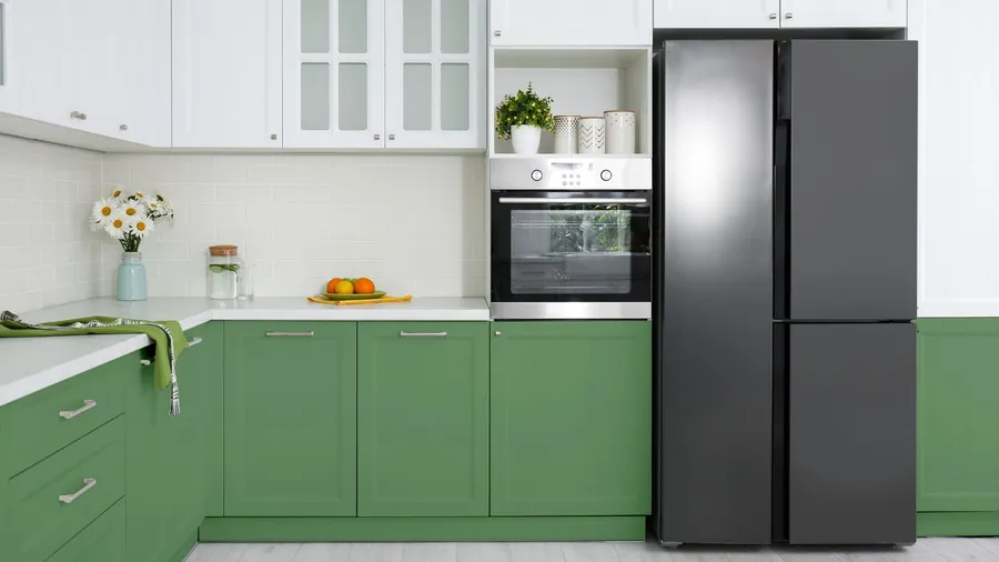 A kitchen interior with white and sage green cabinets, refrigerator, oven, and plants.