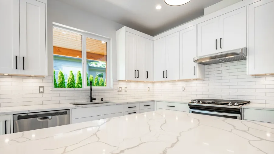 A white kitchen interior with tiles backsplash, crisp white cabinetery, marble countertop, and a window behind the sink.