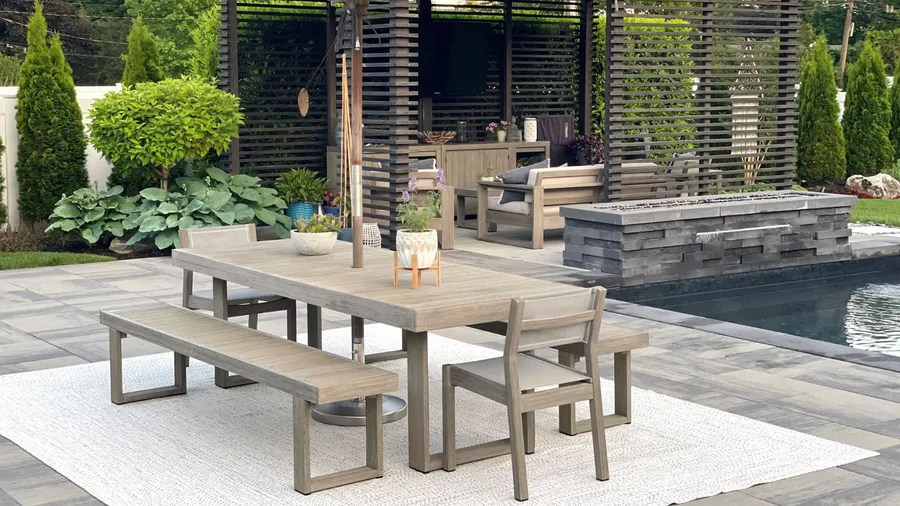An outdoor dining area with a wooden table, chairs, bench with a rug underneath, surrounded by plants.