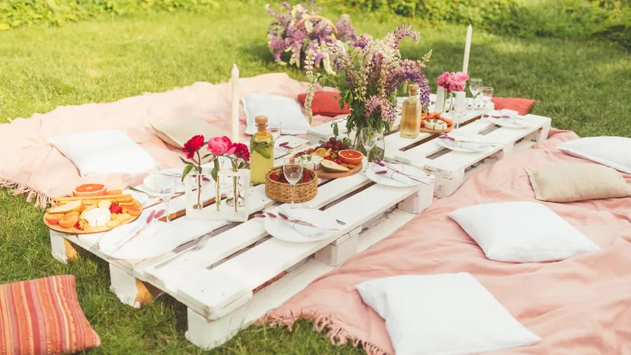 A cozy outdoor dining setup with soft blankets, pillows, and a wooden pallet table set with fresh flowers, candles, and fruits.