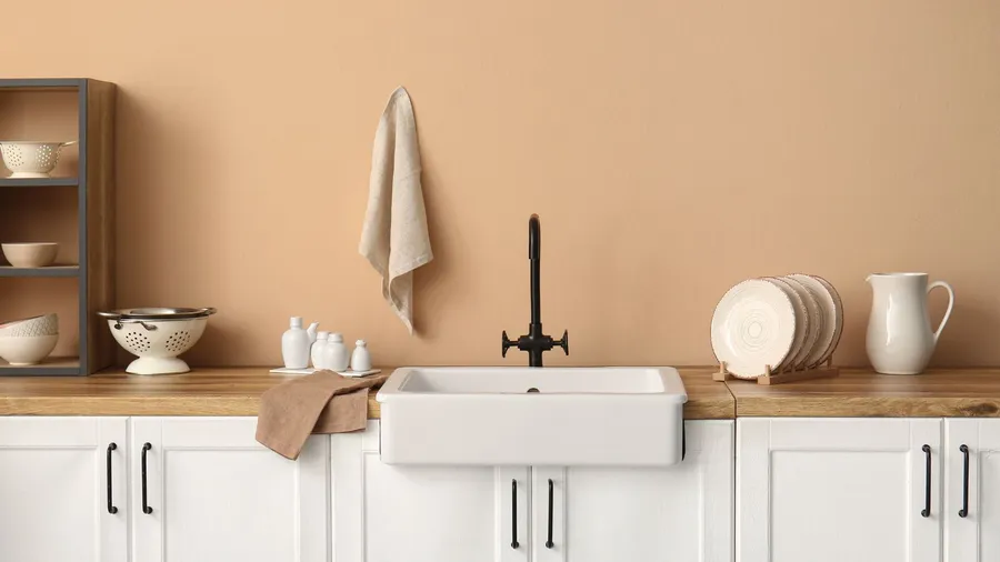 A kitchen interior with a wooden countertop, white cabinets, plates rack, ceramic sink, and towel hanging on a beige wall.