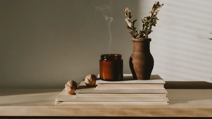 A wooden shelf with a layering of books, candle, nuts, and flower vase.