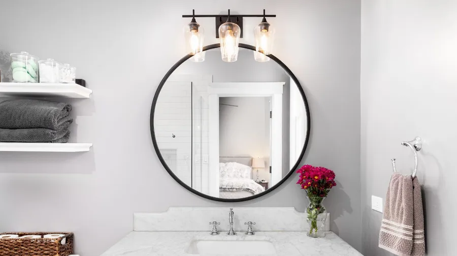 A bathroom interior with a sink, flower vase, towel, floating shelves, and lighting fixture above the mirror.