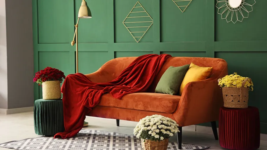 A living room interior with orange sofa, red throw blanket, colorful throw pillows, floor lamp, rug, fresh flowers, and metallic decor accents on the green wall.