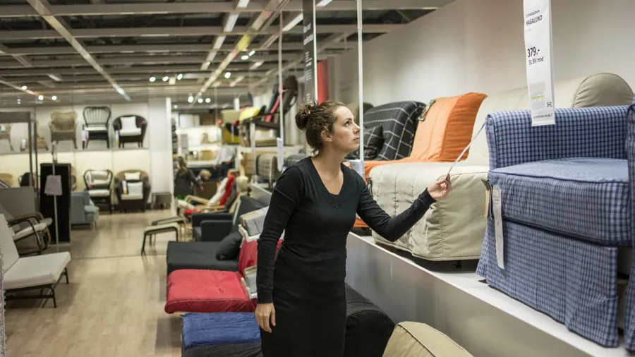 A woman shopping in a furniture store, looking at a bed with orange and gray pillows.
