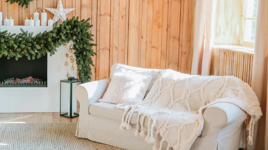 A cozy living room interior with white sofa with fur cushion and knitted throw blanket, rug, lantern, fireplace, and candles and garland on the mantel.