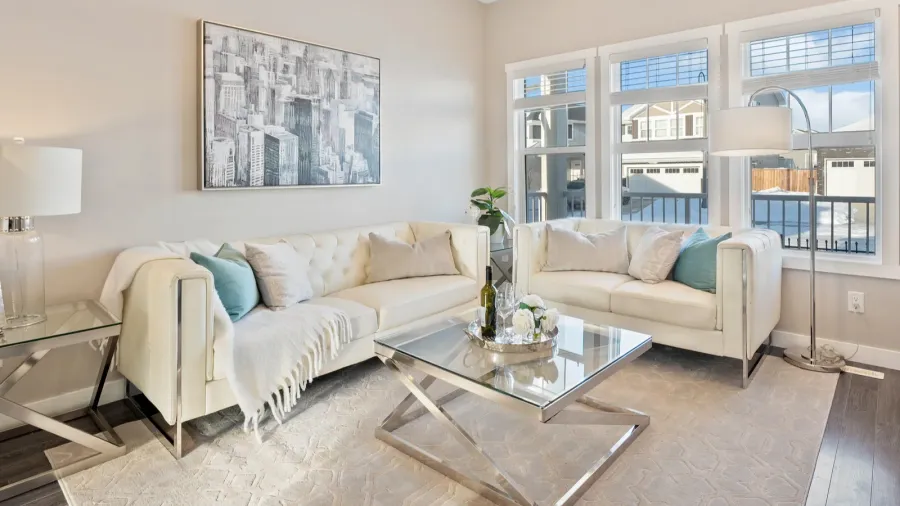 A living room interior with white sofas, glass coffee table, floor lamp, and abstract artwork hanging on the wall.