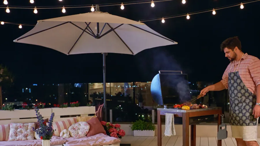 A man grilling barbeque on rooftop patio with cozy seating, potted plants, and illuminated string lights.