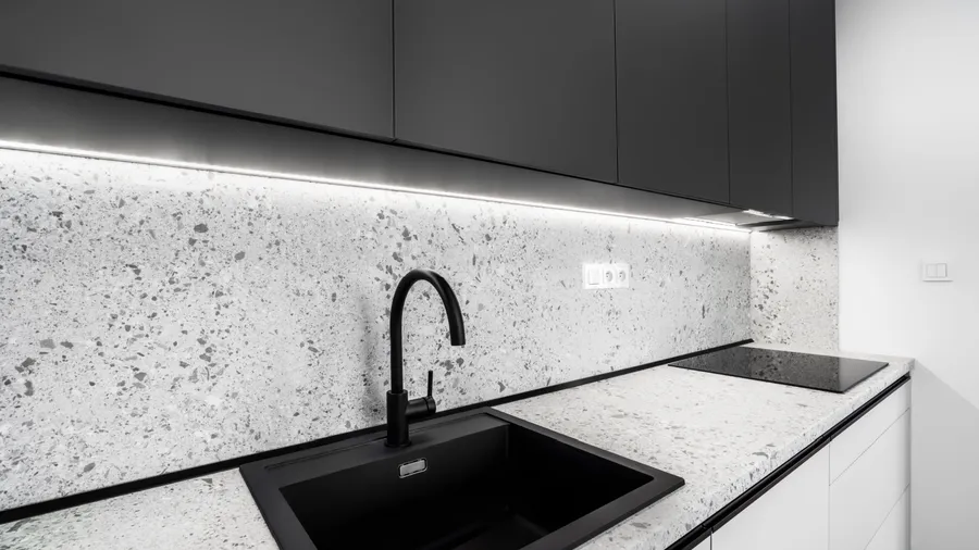A modern kitchen with black cabinets, a terrazzo countertop and backsplash, under-cabinet lighting, a black sink, and an electric cooktop.