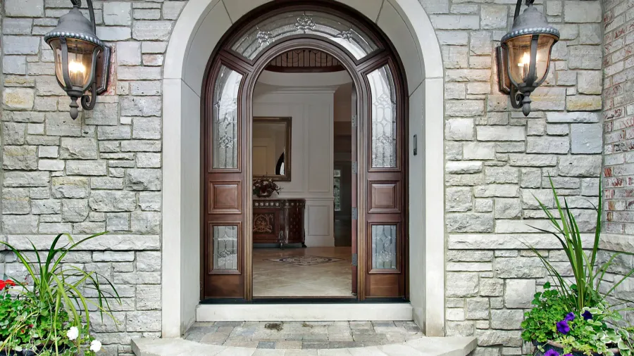 A grand arched wooden front door with decorative glass panels, flanked by two ornate wall lanterns, set within a stone facade with potted plants on either side of the entrance.