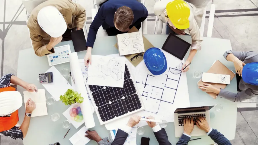 A group of professionals, including architects and engineers, gathered around a table reviewing blueprints, design plans, and a solar panel. Some are wearing hard hats, indicating a construction or design collaboration. The workspace is filled with documents, a laptop, a tablet, and other tools.