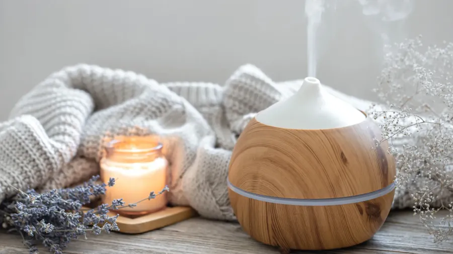 A modern aroma oil diffuser, knitted throw blanket, lavender, and burning candle on a wooden surface.