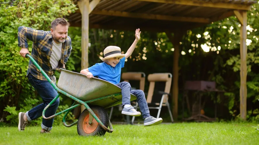 A man pushing a smiling child in a wheelbarrow across a grassy yard with a wooden shelter in the background.