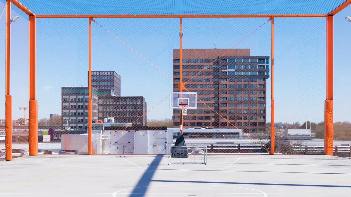 An empty outdoor basketball court with a hoop, surrounded by city buildings under a clear sky.