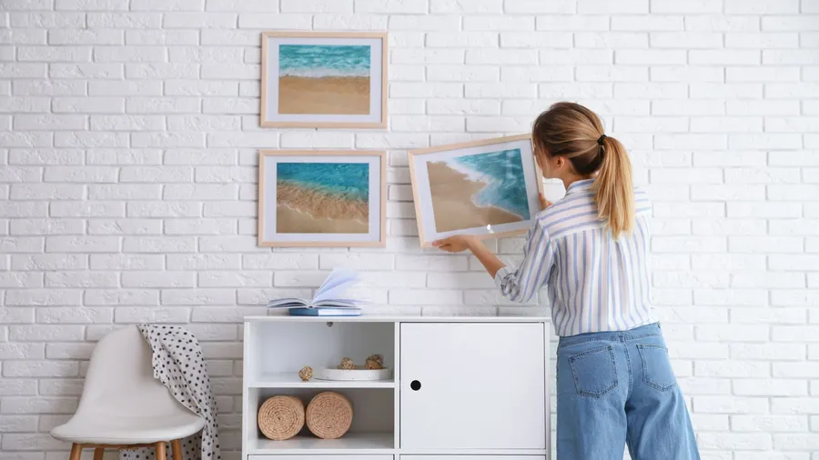 A woman putting a framed picture on a white brick wall.