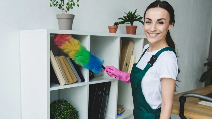 beautiful young cleaner holding duster and smiling at camera while
