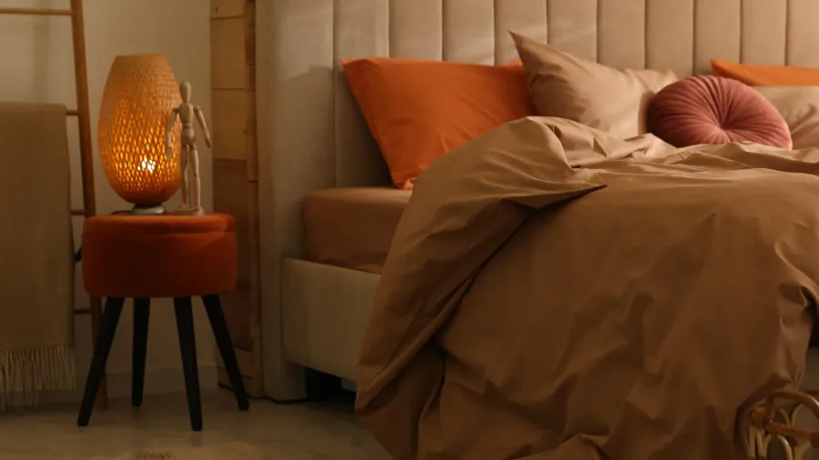 A cozy upholstered bed with earthy color bedding and pillows, and a small stool with lamp and wooden sculpture.