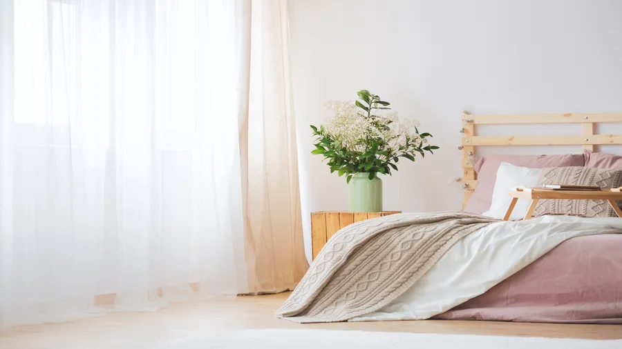 A cozy bedroom with a bed featuring pink bedding and a beige knitted blanket, a wooden nightstand with a vase of white flowers, and sheer white curtains beside a window.