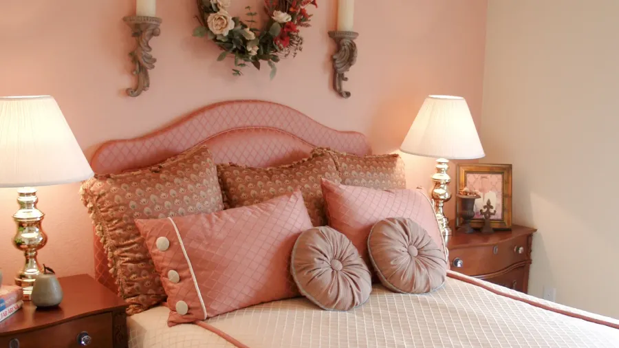 A cozy bedroom with a pink headboard, decorative pillows, bedside lamps, and a wooden dresser adorned with framed art.