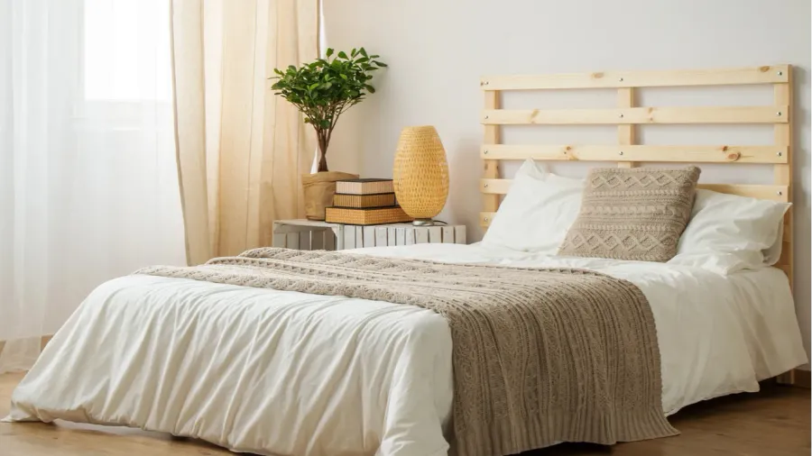 Beige and white minimalistic bedroom interior with cozy bedding, beige throw blanket, side table with lamp, books, and potted plant, and layered sheer curtains on the windows.