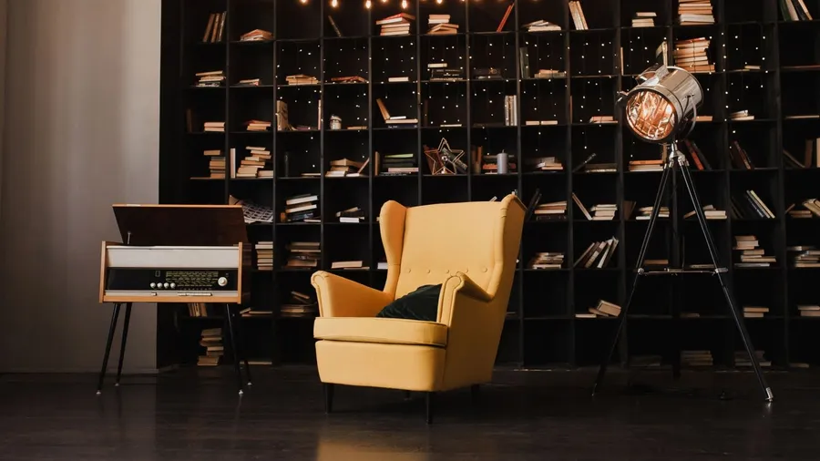 The image shows a cozy room with a yellow armchair, a vintage record player, and a modern lamp. There's a black bookshelf filled with books and decorated with fairy lights.