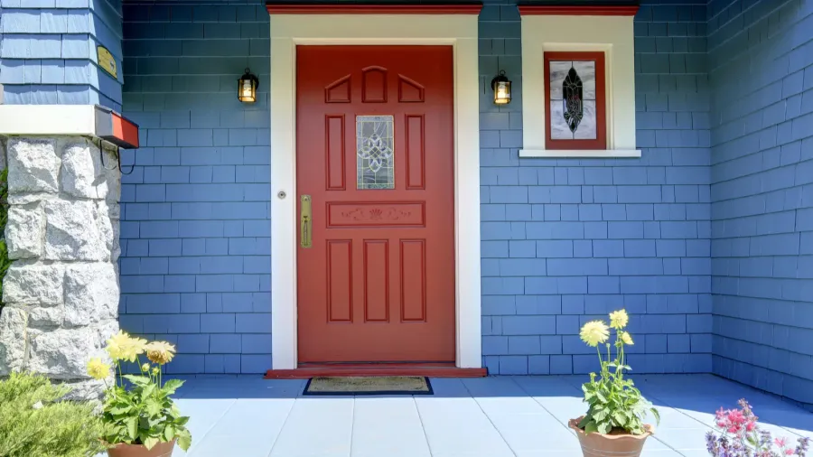 A vibrant red front door on a blue clapboard house, flanked by two outdoor lanterns and potted yellow flowers, with a stone column on the left.