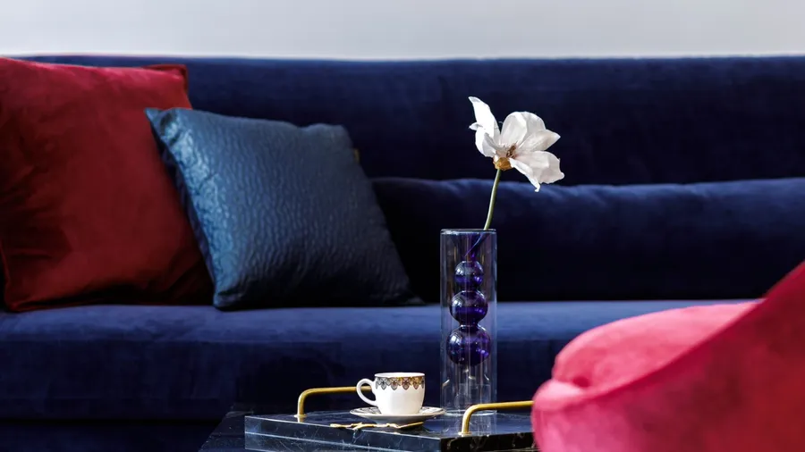 A blue velvet sofa with textured throw pillows and a clear vase with a flower on the table.