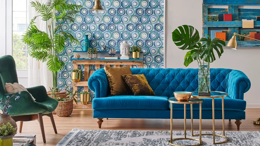 A living room interior with blue wallpaper, blue velvet sofa with cushions, green armchair with woven cushion, and round nesting coffee table.