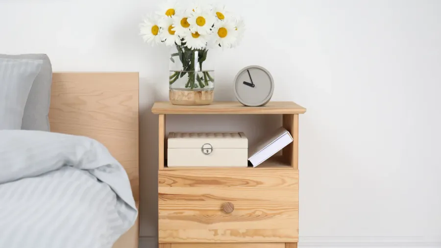 A fresh daisy flower bouquet in a vase on a wooden nightstand with drawer and storage box.