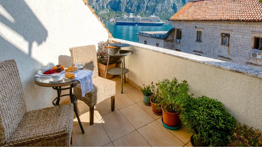 A balcony setup with rattan chairs, table with delicious food, potted plants, and a grill.