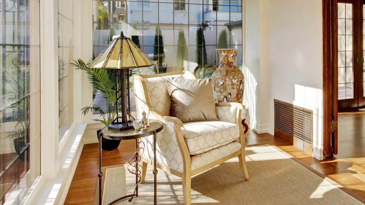 Bright sunlit sitting area with a cozy armchair, table lamp, and decorative vase.