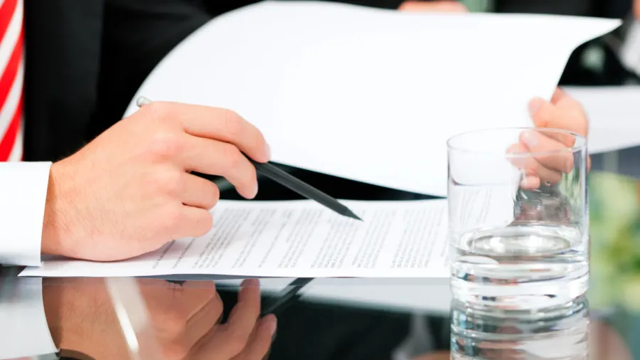 A close-up of a man's hand holding a pen over documents on a table, with a glass of water nearby, suggesting a formal meeting or discussion.