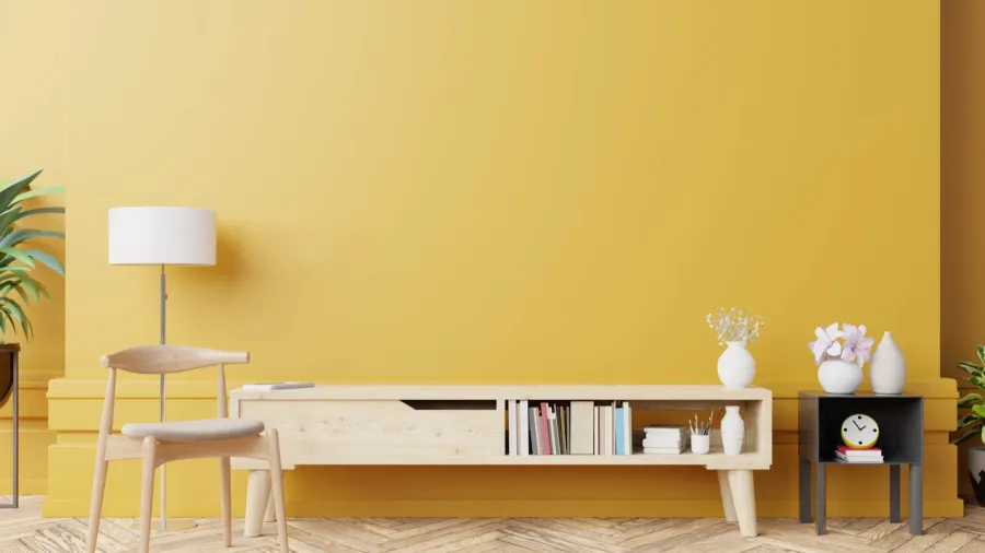 A living room interior with a wooden armchair, floor lamp, plants, and a wooden console on a wooden floor in front of a golden wall.