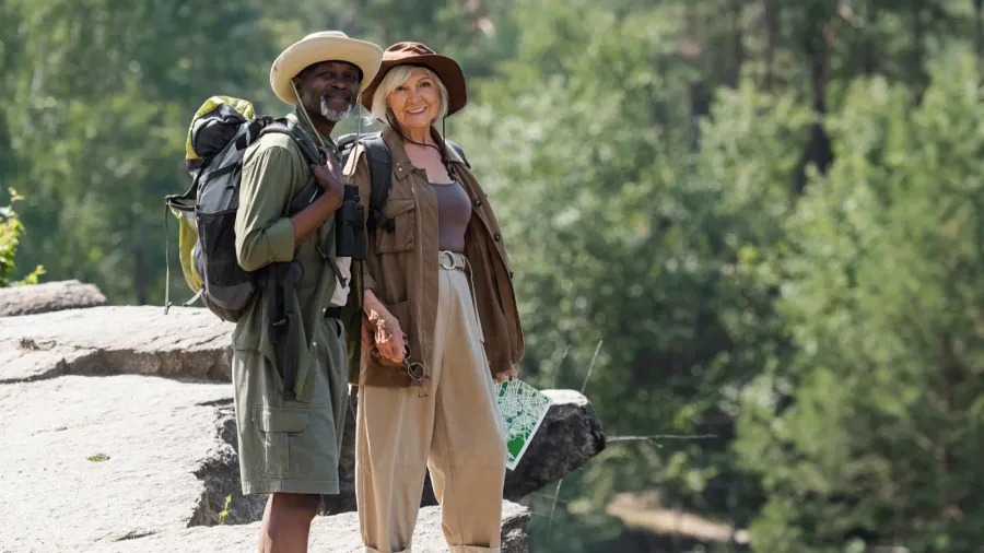 cheerful multiethnic senior travelers with backpacks standing on rock
