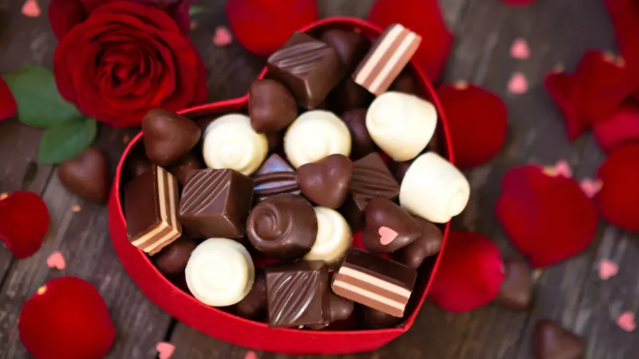 A heart-shaped box of assorted chocolates surrounded by red rose petals.