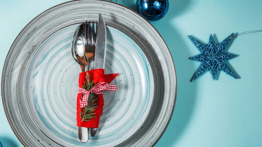 Christmas table place setting on blue background top view. A fork, spoon, and knife wrapped in red cloth and a blue and gray stripes plate with blue Christmas decor scattered around.