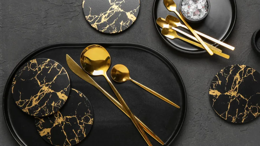 An elegant arrangement of gold utensils on a textured black surface along with marble-patterned coasters.