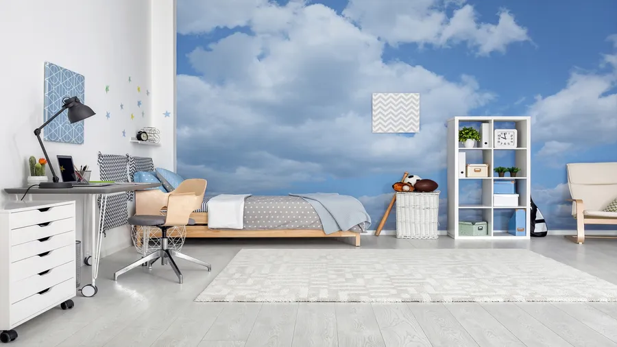 A bedroom interior with bed, study table and chair, storage racks with boxes and decor accents, rug, wall art, and blue sky with clouds wallpaper on the wall.